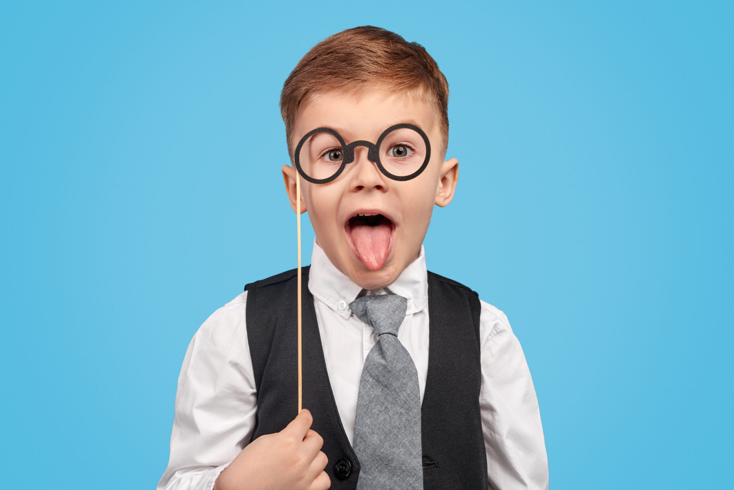 Smart kid in suit holding fake spectacles on stick and showing tongue to camera against blue background
