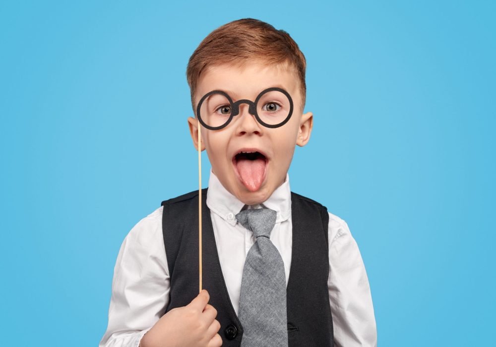 Smart kid in suit holding fake spectacles on stick and showing tongue to camera against blue background